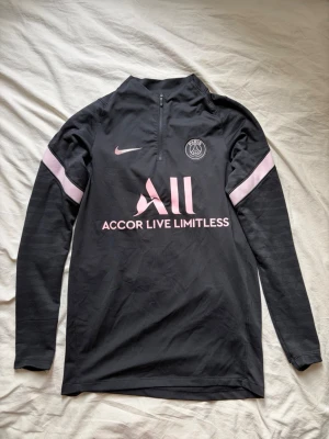 Svart PSG träningströja Nike - Snygg svart långärmad träningströja från PSG med rosa detaljer och halv dragkedja. Tröjan har PSG-logga och Nike Swoosh på bröstet samt sponsortryck fram och bak. Perfekt för fotbollsträning eller chill. Tillverkad i ett lätt och funktionellt material.