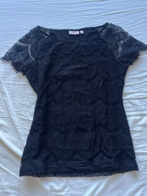 Svart Spetstopp från KappAhl - Snygg svart topp från KappAhl med korta ärmar och genombruten spets. Toppen har rund halsringning 