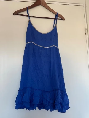 Blå kort klänning från Honolulu - Supersöt blå kort klänning från Honolulu med smala axelband och volang nertill. Klänningen har dekorativa paljettdetaljer på överdelen. Storlek 1 vilket motsvarar S 
