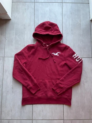 Vinröd hoodie från Hollister - Snygg vinröd hoodie från Hollister med vit logga på bröstet och text på ena ärmen. Klassisk känguruficka framtill och dragsko i huvan. Perfekt för en chill och avslappnad stil.