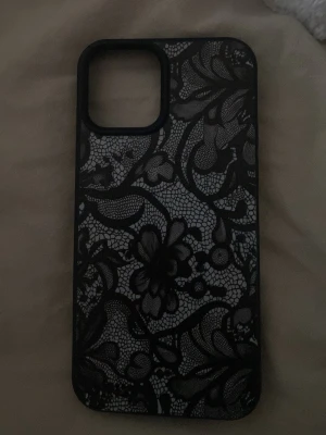 Svart mobilskal med spetsmönster - Snyggt svart mobilskal till en iPhone 12 med detaljerat spetsmönster och blommor. Skalets design är stilren och ger mobilen en unik look. Passar perfekt för dig som vill ha något extra till din telefon. Materialet känns som tålig plast. HAR ALDRIG ANVÄNT!!💕