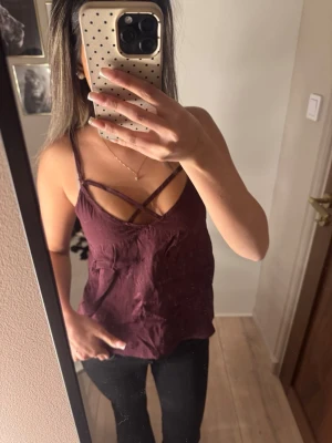 Vinröd linnetopp med korsade band - Säljer en vinröd linnetopp med smala axelband och snygga korsade band framtill. Toppen har en loose passform och är tillverkad i ett mjukt material som känns skönt mot huden. Perfekt för dig som gillar stilrena och trendiga toppar.