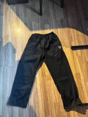 Svarta mjukisbyxor med logga - Säljer ett par svarta mjukisbyxor med vit broderad logga på vänster sida. Byxorna har resår i midjan med dragsko och dragkedjor vid bensluten. Perfekta för chill eller träning. Materialet känns mjukt och bekvämt.