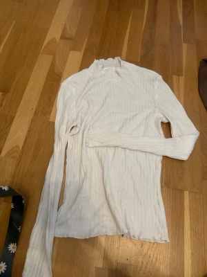 Vit ribbad polotopp med lång ärm - Snygg vit ribbad polotopp med lång ärm och lätt hög krage. Perfekt att styla med jeans eller kjol för en clean look. Tunt och stretchigt material som sitter skönt på kroppen.