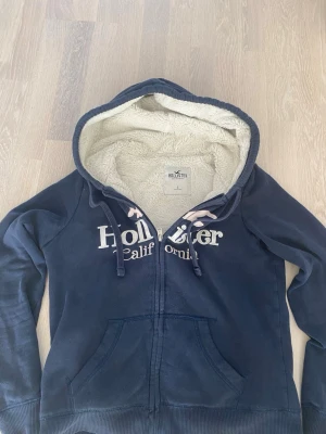 Marinblå hoodie från Hollister - Mysig marinblå hoodie från Hollister med vit fluffig teddyfoder på insidan och stor huva. Dragkedja framtill, två fickor och vit logga över bröstet. Perfekt för chill dagar och när du vill vara extra bekväm.