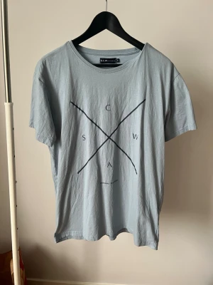 Ljusblå t-shirt från S.C.W - Ljusblå t-shirt från S.C.W med svart grafiskt tryck framtill. Klassisk rund halsringning och korta ärmar. Tillverkad i mjuk bomull för en skön känsla. Perfekt till jeans eller shorts för en avslappnad look.