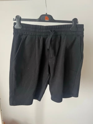 Svarta mjukisshorts med snörning - Svarta mjukisshorts med elastisk midja och snörning. Shortsen har två sidofickor fram och en bakficka. Perfekta för chill dagar eller träning. Tillverkade i mjukt bomullsmaterial för maximal komfort.
