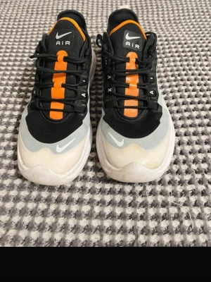 Nike Air Max Axis svart/vit/orange - Nike Air Max Axis sneakers i svart mesh med vita och grå detaljer samt orange inslag på plösen och sulan. Klassisk Nike Swoosh på sidan och AIR-tryck på snörningen. Låg modell med synlig Air Max-dämpning i hälen. Perfekta för en sportig och trendig look.