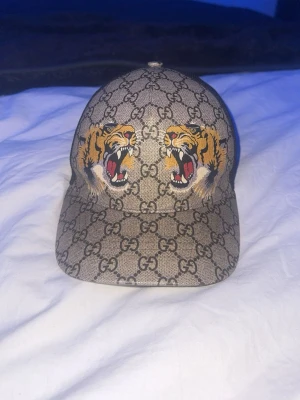 Gucci keps med tigertryck - Snygg keps från Gucci med klassiskt GG-mönster i beige och brunt. Framtill finns två broderade tigrar som ger en cool och unik look. Kepsen har böjd skärm och mesh på baksidan för extra ventilation. Perfekt accessoar för att sticka ut. Kan ochsp byta mot en annan