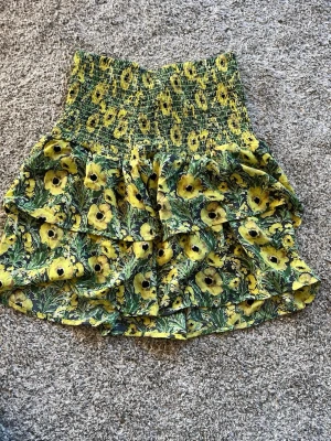 Gul blommig volangkjol med smock - Superfin kort kjol med gult blommönster. Kjolen har smock i midjan och volanger. Använd typ en gång!!