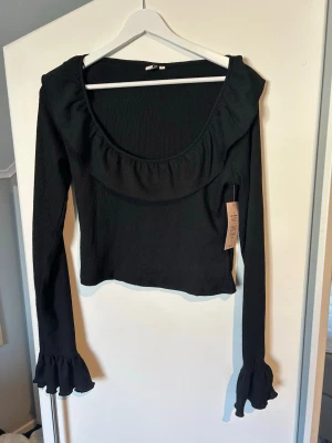 Svart volangtop från Nelly - Trendig svart topp från Nelly med volangdetaljer längs ringningen och vid ärmsluten. Toppen har en djup rundad urringning och långa ärmar. Perfekt för dig som gillar en feminin och stilren look. Aldrig använd och prislappen kvar