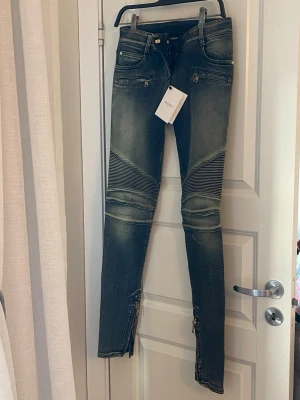 Balmain bikerjeans med dragkedjor - These jeans feature multiple zip pockets, detailed stitching and the recognizable biker panels that make Balmain denim highly sought after.  New with original tags attached.  Retail price: approx. 16,000 SEK (~1,400–1,500 €).  Details 	•	Brand: Balmain Paris 	•	Size: 38 	•	Condition: New with tags 	•	Style: Biker / Moto jeans 	•	Zip pockets 	•	Ribbed biker panels at knees 	•	Zippers at ankles 	•	Premium stretch denim  A statement designer piece from one of the most iconic luxury fashion houses.