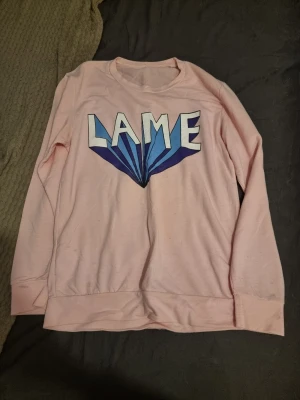 Ljusrosa sweatshirt med tryck - Ljusrosa sweatshirt med stort grafiskt tryck i blått och vitt där det står 'LAME' på bröstet. Tröjan har rund halsringning och långa ärmar. Mjuk och bekväm bomullsblandning, perfekt för en avslappnad stil. Små gråa fläckar på muddarna, kanske går bort med fläck borttagning, men jag har inte testat. Osäker på storlek men den är ganska stor.