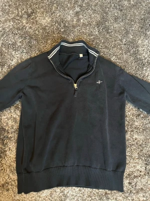 Svart halvzip tröja från Gant - Snygg marinblå tröja från Gant med halvzip och ståkrage. Tröjan har vita ränder på kragen och diskret Gant-logga broderad på bröstet. Tillverkad i mjukt bomullsmaterial och har ribbade muddar vid ärmslut och nederkant.