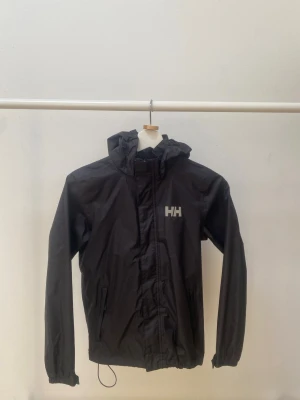 Svart regnjacka från Helly Hansen - Svart regnjacka från Helly Hansen med huva och dragkedja framtill. Jackan har två sidofickor, elastiska muddar och HH-logga på bröstet. Tillverkad i vattenavvisande material, perfekt för regniga dagar.