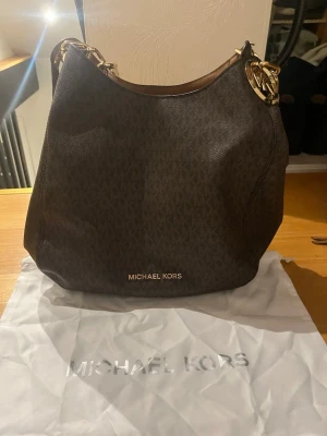 Mörkbrun Michael Kors handväska - Snygg mörkbrun handväska från Michael Kors med guldiga metalldetaljer och klassiskt MK-mönster. Väskan har en mjuk, rundad form och logotypen i guld framtill. Tillverkad i tåligt konstläder och levereras med dustbag och äkthetskort.