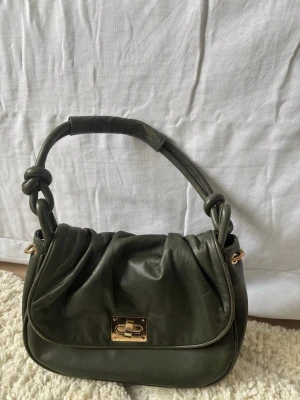 Olivgrön handväska med gulddetaljer - Snygg olivgrön handväska i mjukt skinnliknande material. Väskan har ett unikt handtag med knutdetaljer och guldfärgat vridlås framtill. Perfekt storlek för det viktigaste och en cool accessoar till din outfit. Från märket BTF CPH, nypris ca 1200 kr. Få slitage men endast använd ett fåtal gånger så inget som inte går att ta bort med läderspray/inpregnering. 