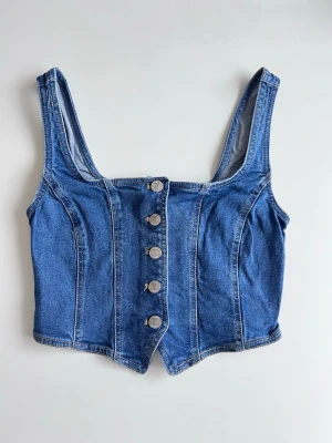 Korsettopp i denim/jeans med knappar framtill, stl S (ny) - Snygg vintage korsettopp stretchig denim. Figurnära passform, ger en snygg y2k-vibe. Så snygg till jeans eller kjol! Storlek 8/S. Inköpt i Australien men blev aldrig använd.