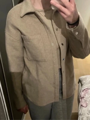 Beige jacka med ficka - Snygg beige vårjacka med klassisk krage och tryckknappar framtill. Jackan har bröstficka på vänster sida och är gjord i ett mjukt, filtliknande material. 