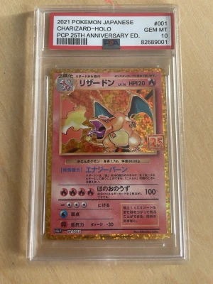Charizard-Holo 25th Anniversary Pokémonkort - Obs ej orginal. Japanskt Charizard-Holo Pokémonkort från 25th Anniversary Edition, graderat PSA 10 GEM MT. Kortet har en glittrig guldram och japansk text, med Charizard i fokus och 25-årslogga. Perfekt för samlare av Pokémonkort och trading cards.