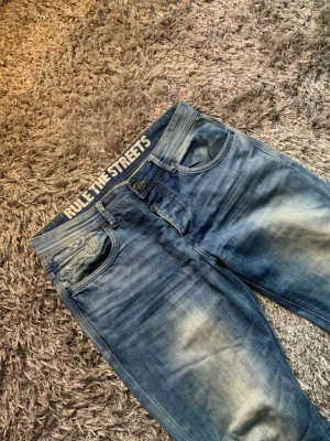 Blå slitna jeans Rule The Streets - Säljer ett par blå jeans med slitningar och tvättade detaljer. Modellen har rak passform och klassisk femficksdesign. På insidan av linningen står det 'RULE THE STREETS' i vitt. Perfekta för en avslappnad streetwear-look.