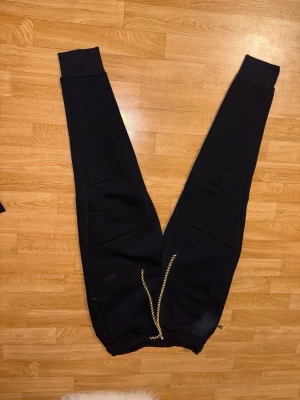 Svarta Nike Tech byxor - Svarta Nike Tech mjukisbyxor med breda muddar vid benslut och midja. Byxorna har snörning i midjan med svart och vit mönstrad dragsko samt fickor på sidorna. Perfekta för en avslappnad stil.