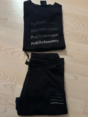 Svart Peak Performance set - Mysigt svart loungewear-set från Peak Performance med matchande sweatshirt och byxor. Tröjan har flera logotyper tryckta framtill i olika nyanser av svart och vitt. Byxorna har resår och dragsko i midjan samt logga på benet. Perfekt för chill dagar hemma.