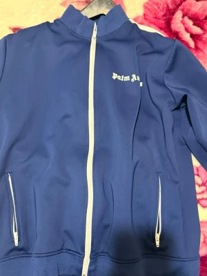 Blå tracksuit från Palm Angels - Snygg blå tracksuit från Palm Angels med vita detaljer. Jackan har vit dragkedja framtill, två fickor med dragkedja och vit textlogga på bröstet. Byxorna har vita ränder längs sidorna. Perfekt för en sportig och trendig look.