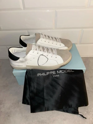 Philippe Model vita sneakers - Stilrena vita sneakers Philippe Model med grå mockadetaljer på tån och tungan samt svart hälparti. Skorna har en klassisk låg silhuett, vita skosnören och diskret logga på sidan. Tillverkade i läder och mocka för en lyxig känsla.