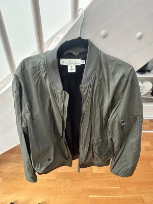 Olivgrön bomberjacka från L.O.G.G. By H&M - Snygg olivgrön bomberjacka från L.O.G.G. by H&M med klassisk ribbad krage och muddar. Jackan har dragkedja framtill, fickor med knapp och en ärmficka med dragkedja. Tillverkad i ett lätt, glansigt material som ger en cool streetkänsla.