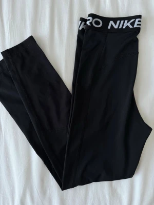 Svarta Nike Pro leggings - Svarta Nike Pro leggings med bred resår i midjan där det står 'NIKE PRO' i vitt. Tight passform och stretchigt material som är perfekt för träning eller chill. Klassisk och sportig look med diskreta sömmar.