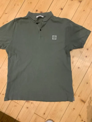 Grön pikétröja från Stone Island - Snygg grön pikétröja från Stone Island med klassisk krage och två knappar framtill. Tröjan har en diskret logotyp på bröstet och är tillverkad i bomull för en bekväm känsla. Perfekt till alla stilar! Bara att höra av dig vid minsta fråga 