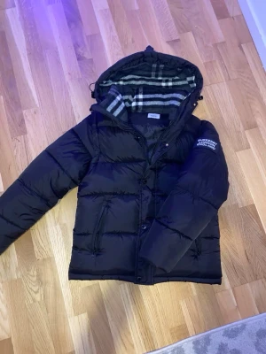 Svart pufferjacka från Burberry - Säljer en svart pufferjacka från Burberry med avtagbar huva och klassiskt rutigt foder. Jackan har dragkedja och knappar framtill, två fickor och texten 'Burberry London England' på ärmen. Perfekt för kalla vinterdagar och riktigt snygg streetstil.