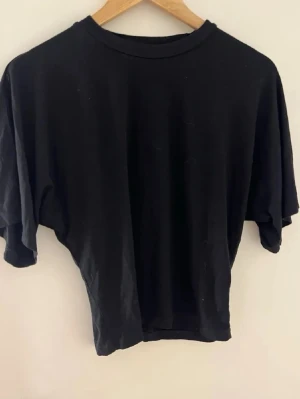 Svart croppad t-shirt - En stilren svart t-shirt med croppad passform och korta ärmar. T-shirten har en klassisk rund halsringning och är tillverkad i mjuk bomull som känns skön mot huden. Perfekt att matcha med kjol eller jeans för en clean look.