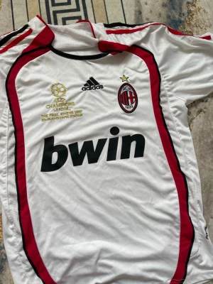 AC Milan retro tröja  - Säljer en vit AC Milan matchtröja från Adidas med röda och svarta detaljer. Tröjan har Champions League-brodyr, klubbmärke och Bwin-tryck på bröstet. Tillverkad i lätt och ventilerande material, perfekt för fotboll eller samling.