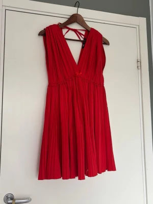 Röd plisserad kortklänning med knyt - Superfin röd kortklänning med plisserat tyg och v-ringning. Klänningen har knytband i ryggen och en liten rosett framtill. 
