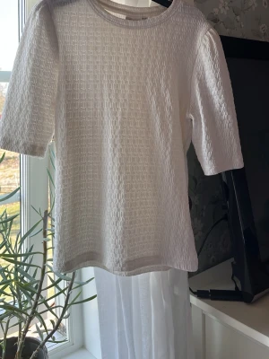 Vit strukturerad topp med halvärm - Snygg vit topp med strukturerat tyg och rund hals. Modellen har halvärm och rak passform, vilket ger en clean och modern look. Perfekt att styla till jeans eller kjol för en fräsch outfit.