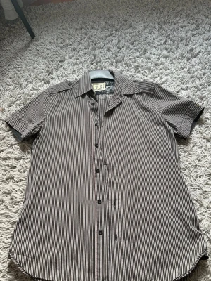 Randig kortärmad skjorta från Diesel - Snygg kortärmad skjorta från Diesel med smala svarta och vita ränder. Skjortan har klassisk krage, knappar framtill och coolt tryck på insidan av kragen. Perfekt för dig som gillar streetwear och vill sticka ut lite extra.