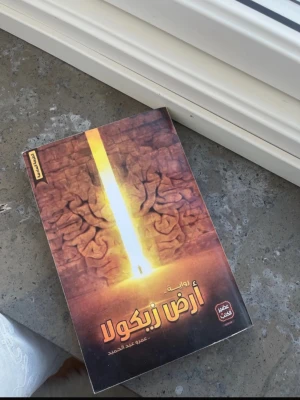 Arabiska romanen 'Ard Zibikola' - En skönlitterär bok på arabiska med titeln 'Ard Zibikola'. Omslaget har ett mystiskt motiv i bruna och gula toner med ett ljusstråk i mitten och mönstrade stenväggar. Perfekt för dig som gillar äventyr och fantasy.