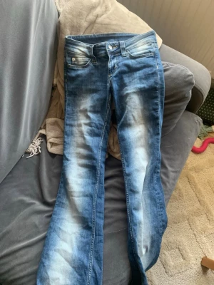Blå bootcut jeans med slitningar - Säljer ett par blå bootcut jeans med tydliga slitningar och ljusa tvättade partier framtill. Jeansen har låg midja och klassisk femficksdesign. Materialet är denim i bomull. Perfekta för en avslappnad och trendig look.