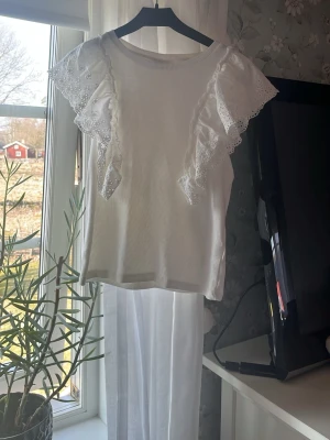 Vit blus med spetsvolang - Supersöt vit blus med ribbad struktur och stora spetsvolanger över axlarna. Blusen är ärmlös och har rund halsringning. Perfekt för dig som gillar romantiska detaljer och en ljus, fräsch look.