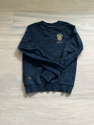 Svart sweatshirt Morris R Club - Svart sweatshirt från Morris R Club med broderad logga och brittisk flagga på bröstet. Tröjan har rund halsringning, långa ärmar och ribbade muddar vid ärmslut och nederkant. Perfekt för en avslappnad och stilren look.