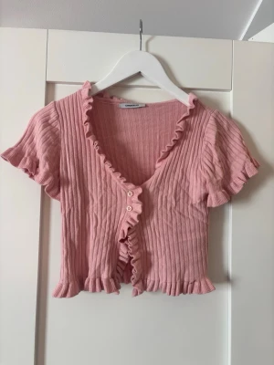 Rosa ribbad croppad topp med volang - Supersöt rosa ribbad topp från Chiquelle med korta ärmar, volangkant längs v-ringningen och nederkanten samt knappar framtill. Croppad modell som ger en trendig look och passar perfekt till höga jeans eller kjol.