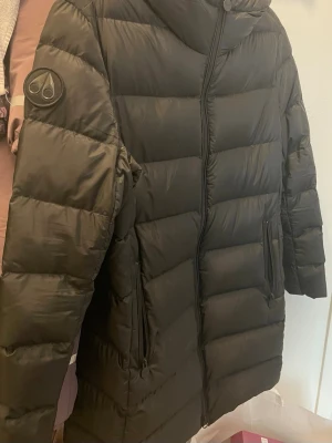 Svart dunjacka Moose Knuckles - Säljer en svart dunjacka från Moose Knuckles med hög krage och dragkedja framtill. Jackan har två stora fickor med dragkedja och en snygg logga på ärmen. Perfekt för kalla vinterdagar och riktigt stilren look.