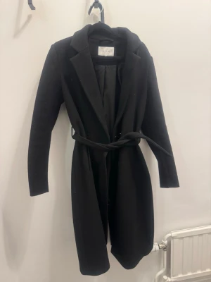 Svart kappa med skärp från VILA - Stilren svart kappa från VILA med klassisk krage och knytbart skärp i midjan. Kappan har lång ärm och rak passform, perfekt för en trendig look. Materialet är mjukt och ger en snygg siluett.