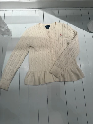 Vit kabelstickad kofta från Polo Ralph Lauren - Priset går att diskutera till lägre!❣️Kabelstickad kofta i krämvitt från Polo Ralph Lauren med volangkant nedtill och rund hals. Långa ärmar och knäppning framtill. Klassisk röd logga broderad på bröstet. Perfekt för en klassisk look och enkel att matcha. Knappen längst ner är borta men det är inget man ser. Storleken är 12-14 år (L) men passar på en xs i vuxen storlek också