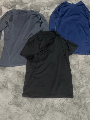 Träningsset med tre sporttröjor - Säljer ett set med tre sporttröjor i funktionsmaterial. En långärmad grå, en långärmad blå och en kortärmad svart. Perfekta för träning eller löpning, med bekväm passform och snabbtorkande tyg.