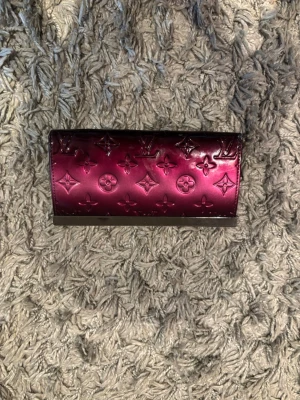 L.V plånbok i lack - Säljer en glansig lila plånbok från L.V i lackat läder med det klassiska monogrammönstret. Plånboken har flera fack, dragkedja och plats för kort. Perfekt accessoar för dig som gillar lyxiga detaljer och snygg design.