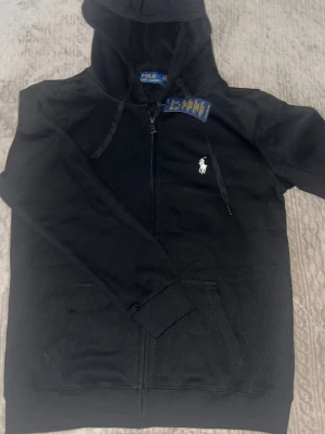 Svart hoodie från Polo Ralph Lauren - Snygg svart hoodie från Polo Ralph Lauren med dragkedja framtill och klassisk vit broderad logga på bröstet. Huva med dragsko och känguruficka. Tillverkad i mjukt bomullsmaterial som är skönt att ha på sig. Perfekt för en avslappnad och stilren look.