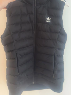 Svart dunväst från Adidas - Snygg svart dunväst från Adidas med dragkedja framtill och två praktiska fickor med dragkedja. Västen har Adidas klassiska logga på bröstet och quiltad design som ger en sportig look. Perfekt för lager-på-lager och streetwear.
